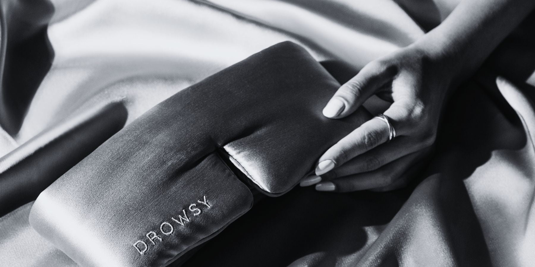 A hand with manicured nails holds a silky DROWSY branded sleep mask on a smooth satin fabric background.