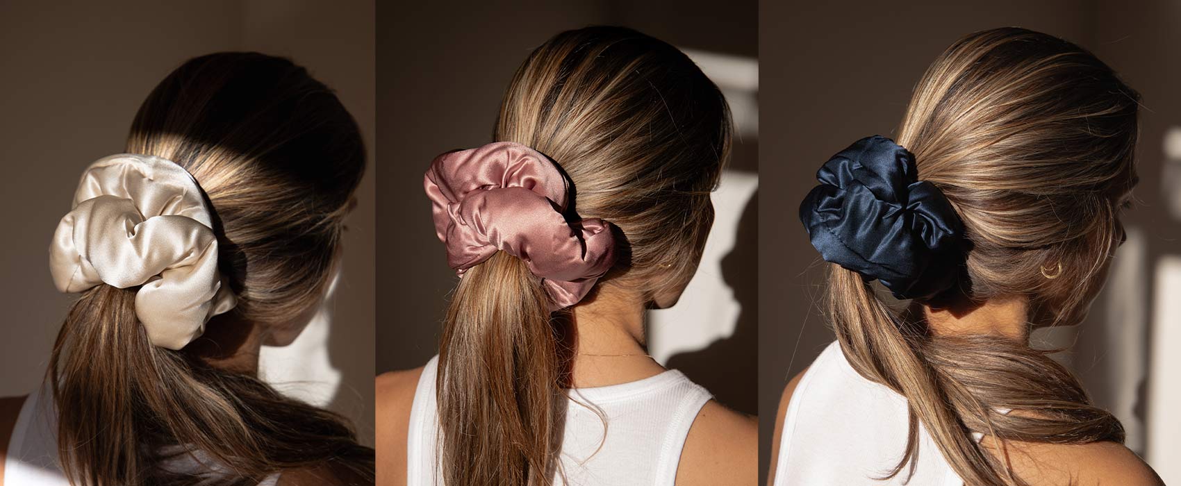 Three women with light brown, wavy hair are shown from behind, each wearing a different large satin scrunchie—cream, pink, and navy—holding their low ponytails, in soft natural light.