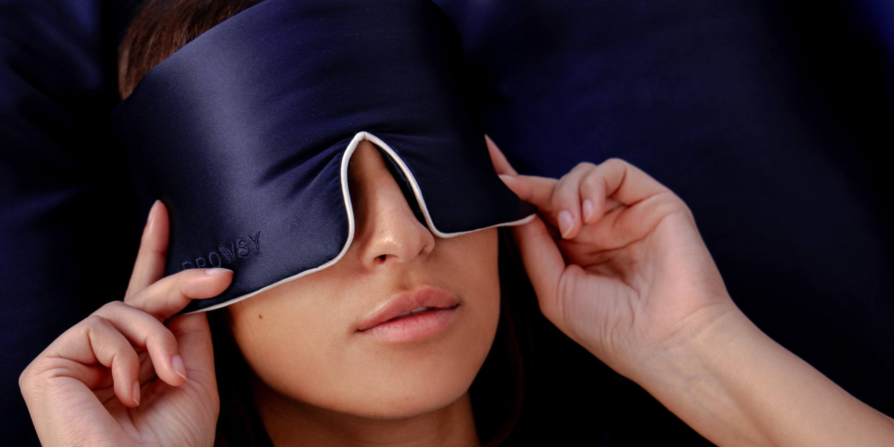 A woman reclines while adjusting a dark blue sleep mask over her eyes, with her hands gently touching the mask and her lips slightly parted.