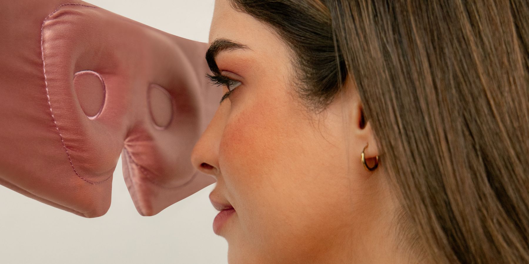 A woman with brown hair and gold hoop earrings faces a large, plush, pink nose, almost touching it, against a plain background.
