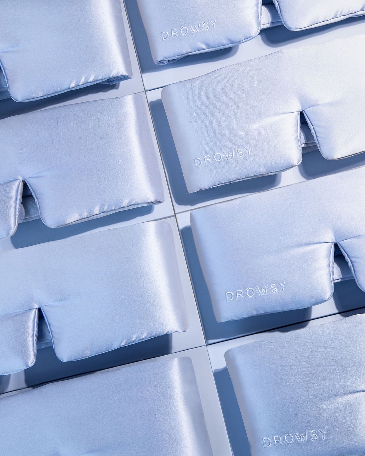 A flat lay of multiple light blue satin sleep masks arranged neatly in rows. The word DROWSY is embroidered on each mask in white lettering.