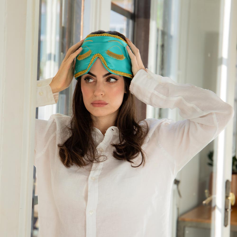 A woman with long brown hair, wearing a white button-up shirt, adjusts the turquoise silk Breakfast at Drowsy sleep mask on her head as she stands indoors, gazing thoughtfully to the side.