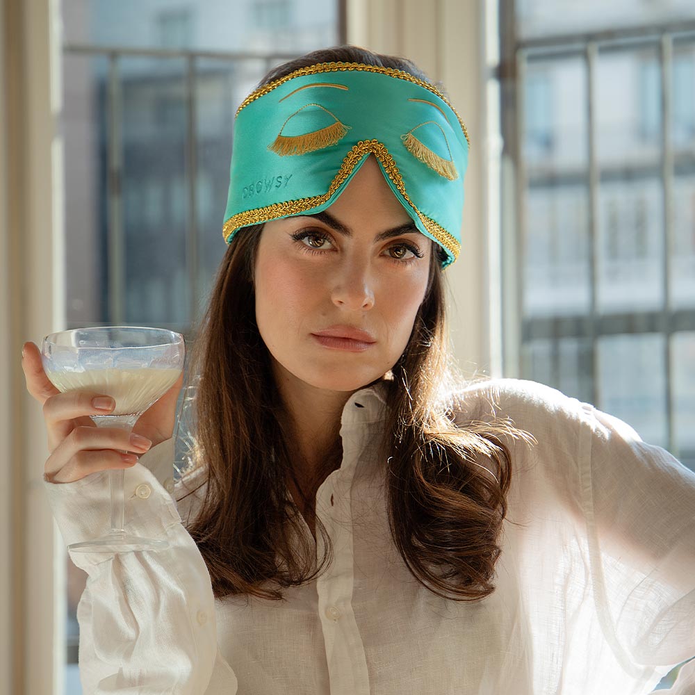 A woman in a white shirt wears the Breakfast at Drowsy turquoise mulberry silk sleep mask with gold trim and embroidered eyes, holding a light drink, standing before tall windows overlooking city buildings.