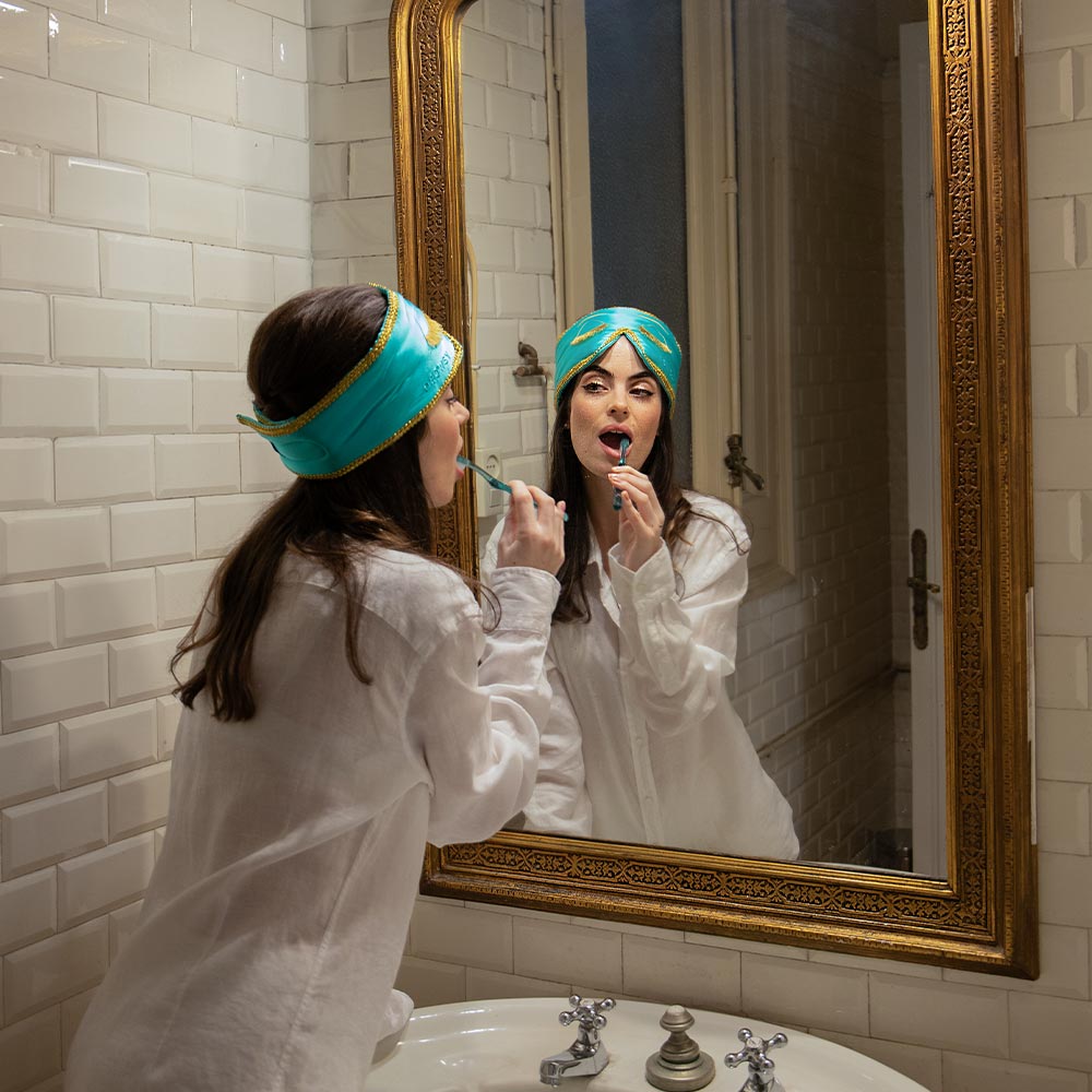 A woman in a blue headband and white shirt applies lipstick before a large ornate mirror in a white-tiled bathroom, her Breakfast at Drowsy mulberry silk sleep mask resting on the counter beside her.