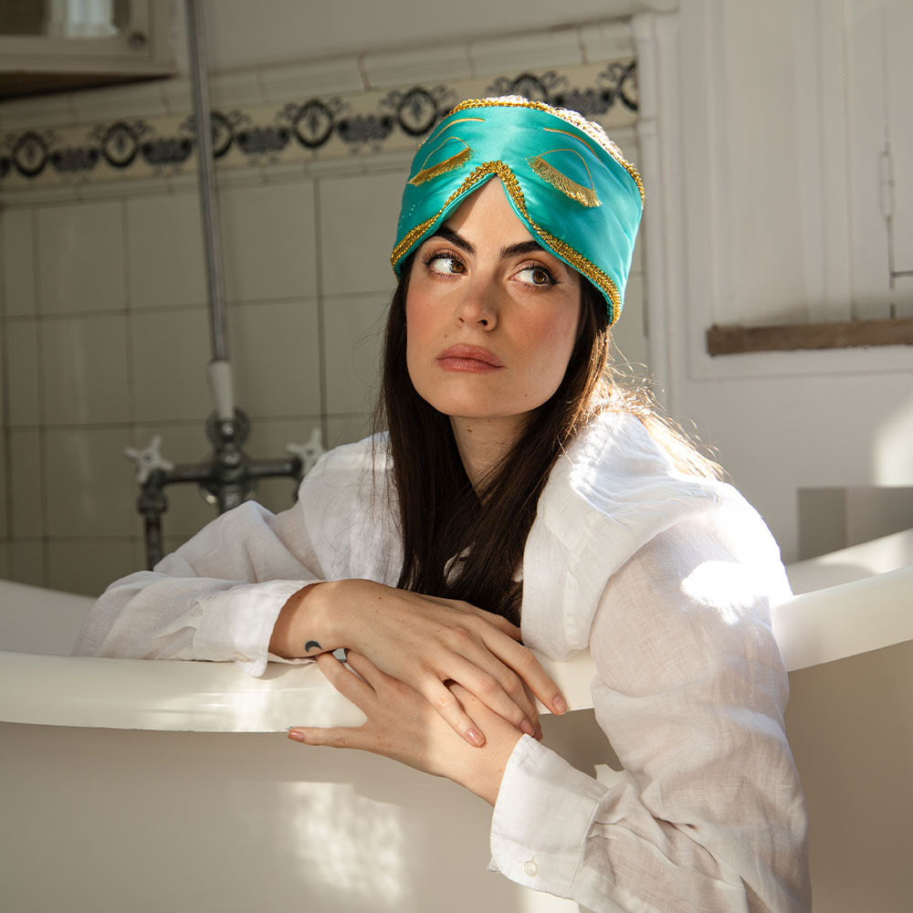 A woman in a white shirt sits thoughtfully in a sunlit bathtub with the turquoise and gold-trimmed Breakfast at Drowsy mulberry silk sleep mask pushed up on her head.