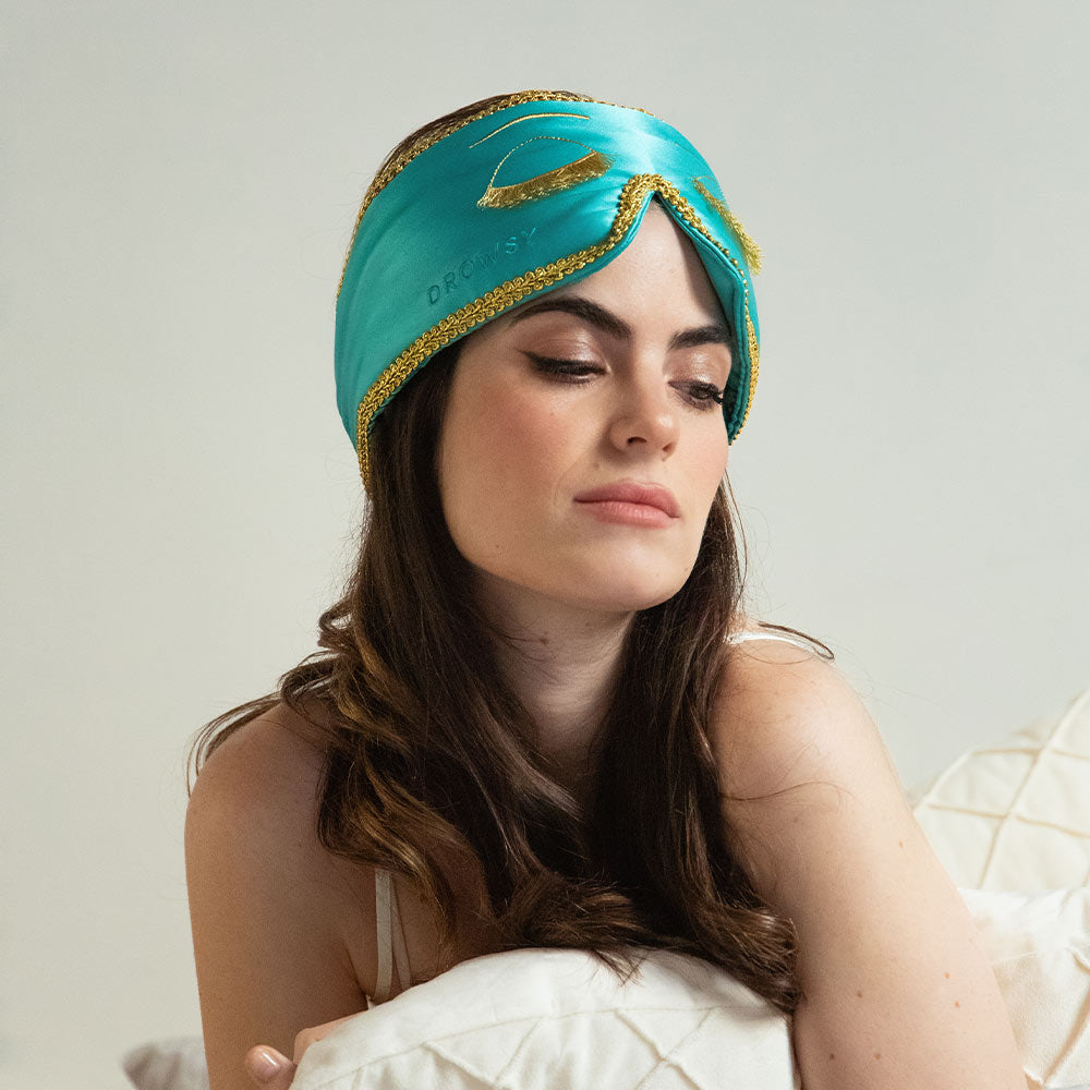 A woman with long brown hair sits on a bed, wearing the Breakfast at Drowsy teal mulberry silk sleep mask with gold trim, gazing downward with a relaxed expression against a softly lit neutral background.