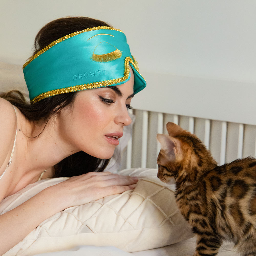 A woman wearing the Breakfast at Drowsy turquoise silk sleep mask with gold trim lies on a bed, gazing at a brown spotted kitten near her pillow.