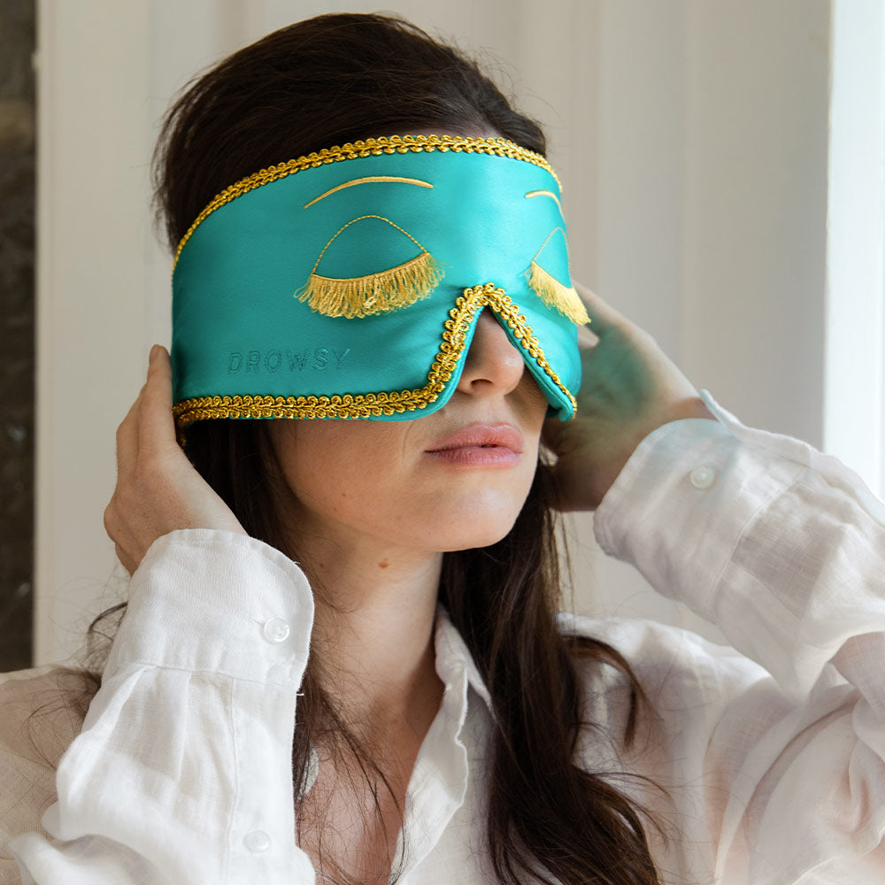 A woman wearing a turquoise sleep mask with gold fringe and trim, designed to look like closed eyes with lashes, adjusts the mask while dressed in a white shirt.