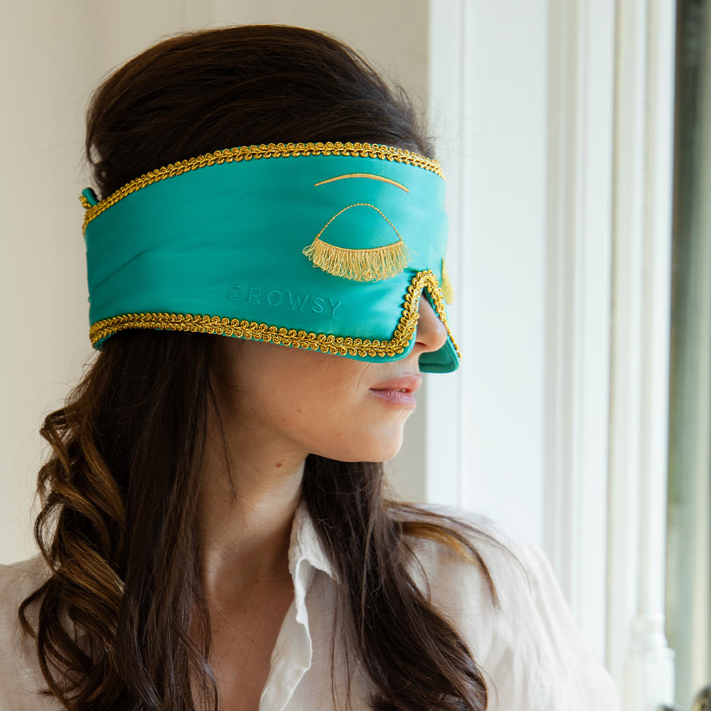 A woman with wavy brown hair stands sideways by a bright window, wearing the Breakfast at Drowsy teal silk sleep mask with gold trim and a gold eyelash design.
