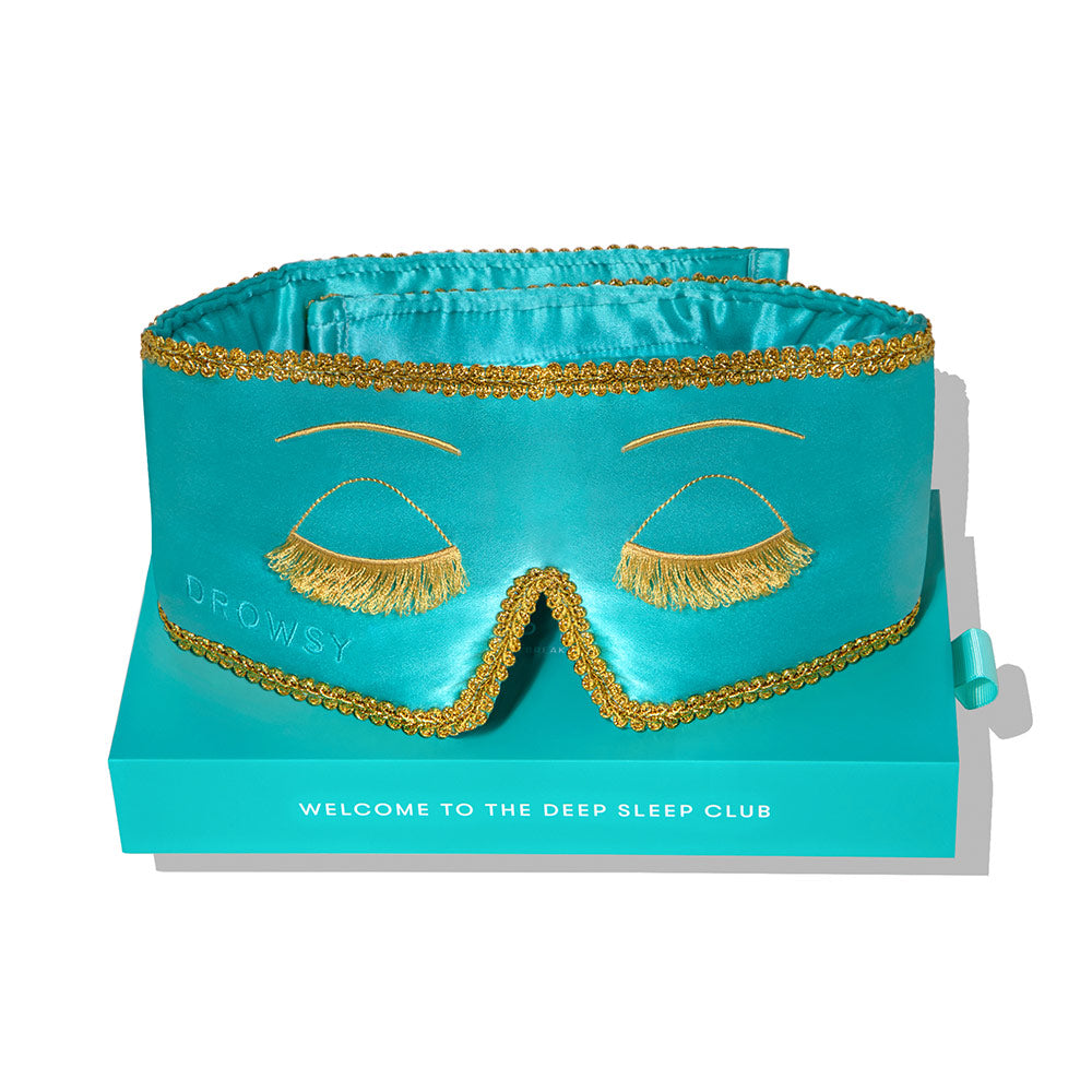 The “Breakfast at Drowsy” mulberry silk sleep mask in teal features gold fringe lashes, embroidered closed eyes, and gold trim. It comes in a matching “Welcome to the Deep Sleep Club” box and has a blackout design with “Drowsy” on the side.