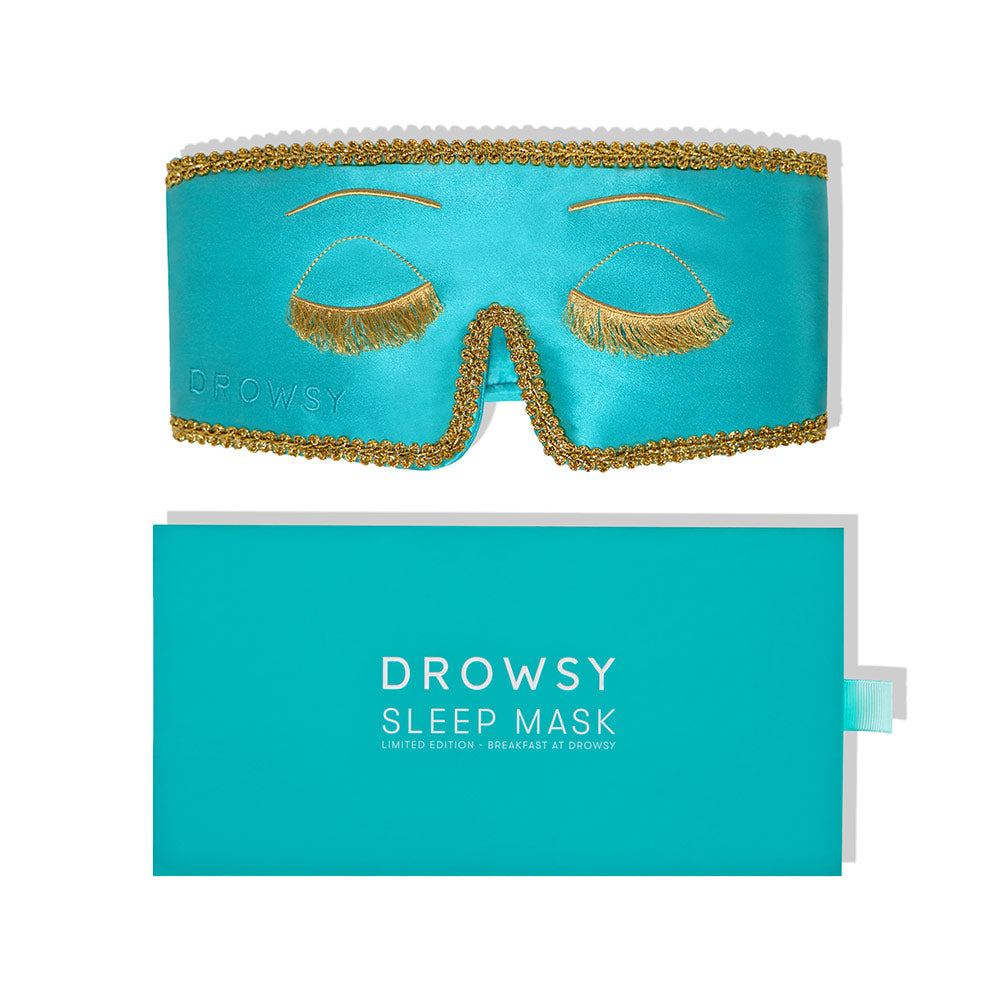 A turquoise mulberry silk sleep mask, Breakfast at Drowsy, with gold trim and embroidered closed eyes with gold lashes, sits above a matching turquoise box. Its blackout design delivers luxurious, uninterrupted rest.
