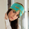 A woman with long brown hair and a relaxed expression leans against a white wall, wearing the Breakfast at Drowsy turquoise silk sleep mask with gold trim and tassel eyelash details pushed up on her forehead.