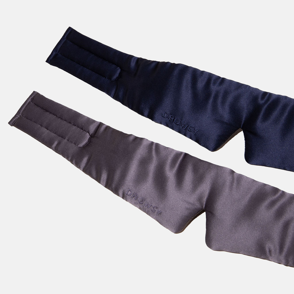 The Lovers Pack features two padded, satin-finish sleep masks in navy blue and gray, each with a wraparound design, Velcro straps, and “DROWSY” embroidered on the front. Displayed on a white background.