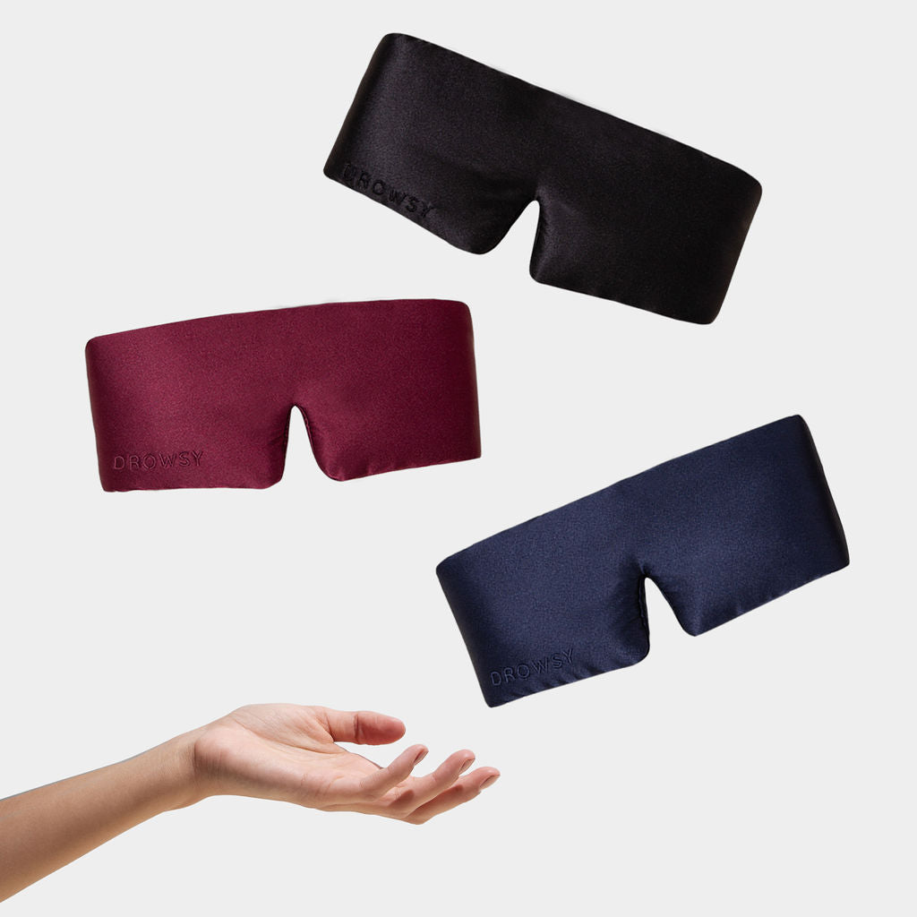 A hand reaches upward beneath three floating Besties Pack sleep masks in black, burgundy, and navy against a plain white background.