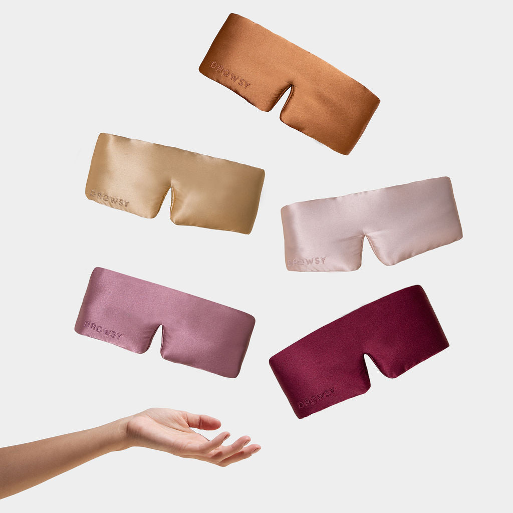 A hand reaches toward the Family Pack of five satin sleep masks in brown, gold, pink, mauve, and burgundy, displayed against a plain white background.