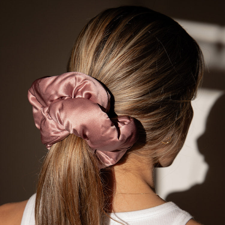 A woman with light brown hair pulled into a low ponytail, secured with a large, shiny mauve satin scrunchie. She is wearing a white sleeveless top, and the photo is taken from behind in natural light.