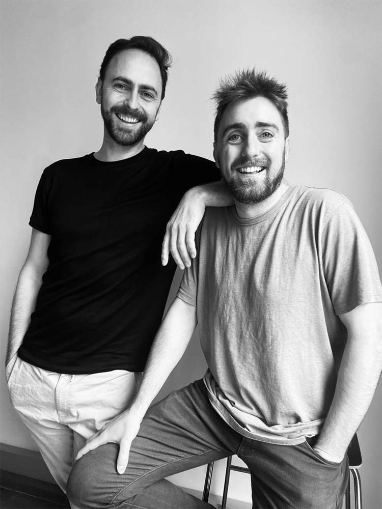 Two men smiling at the camera. One stands with his hands in his pockets, wearing a black T-shirt. The other sits on a stool, leaning on the first mans shoulder, wearing a light gray T-shirt.
