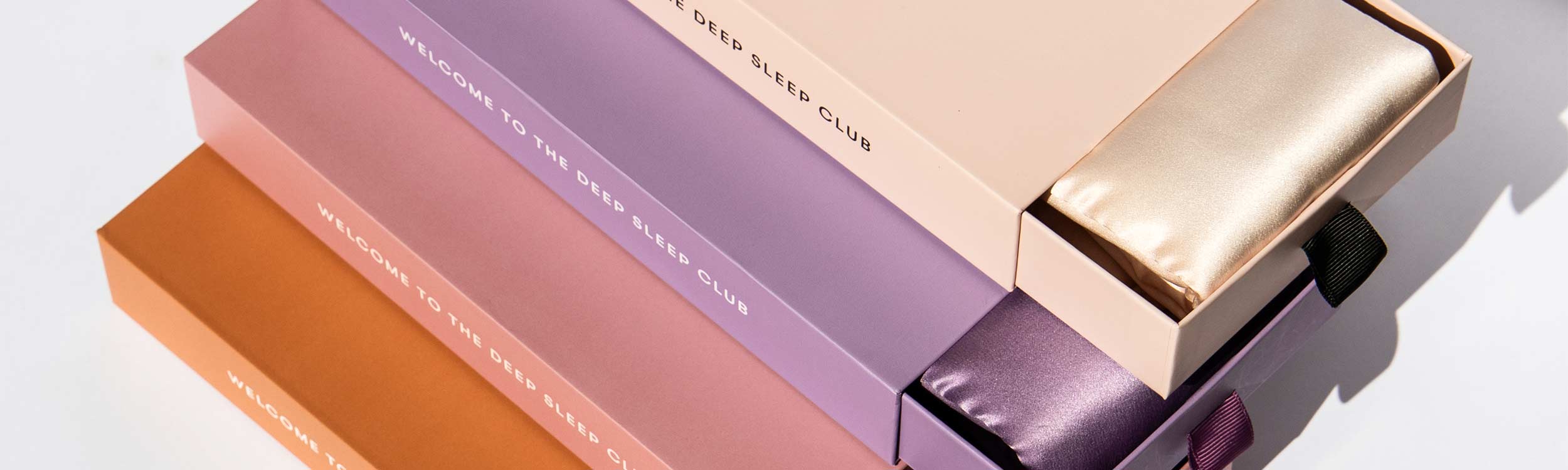 Four pastel-colored boxes, each with “Welcome to the Deep Sleep Club” printed on them, are stacked together. Two boxes are slightly open, revealing silk sleep masks inside.