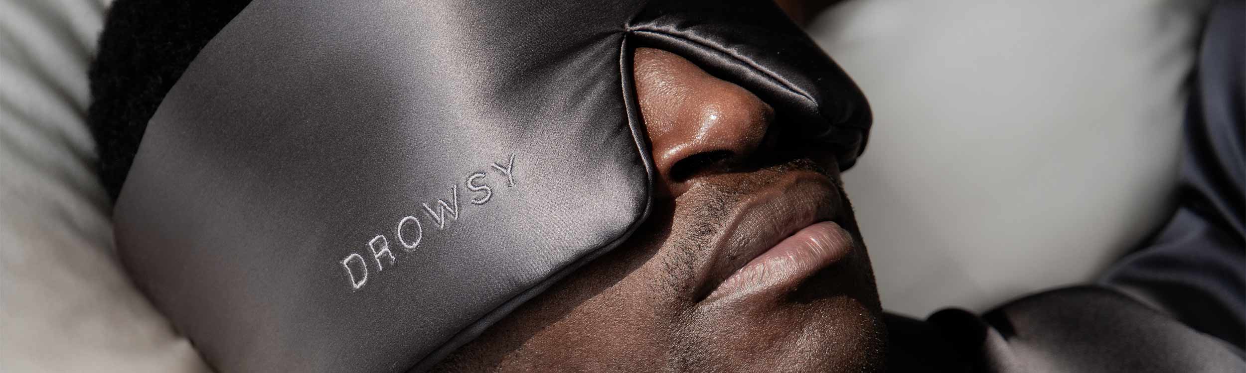 A person wearing a large, silky black sleep mask with the word DROWSY embroidered on it, lying down with eyes covered, suggesting rest or sleep.