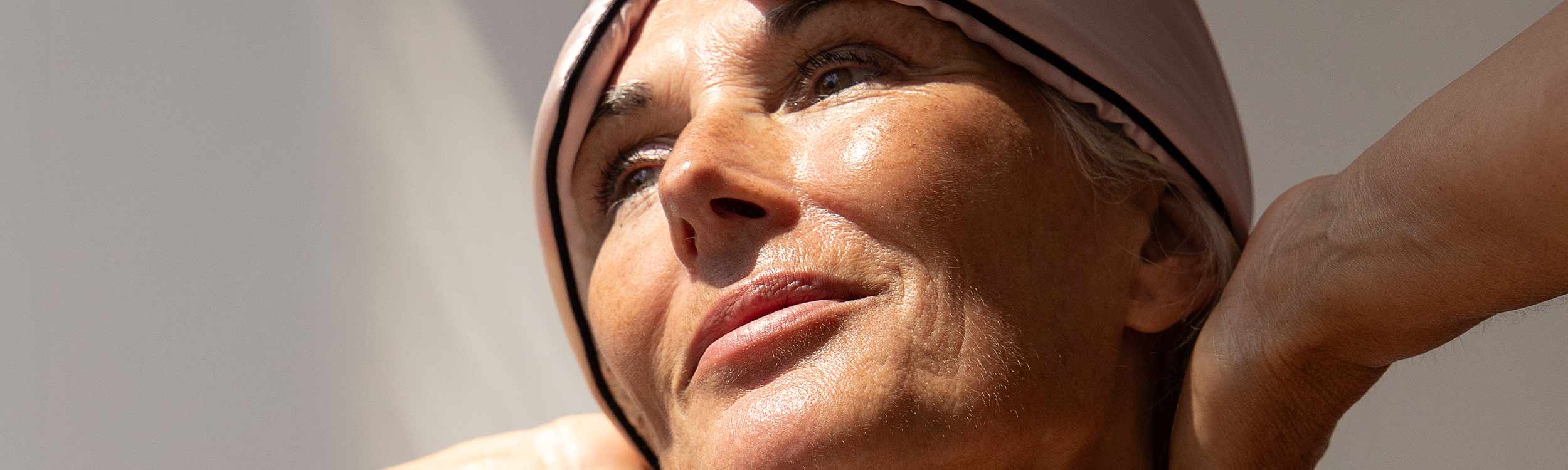 Close-up of an older woman wearing a light pink headscarf, looking relaxed and content with sunlight highlighting her face.