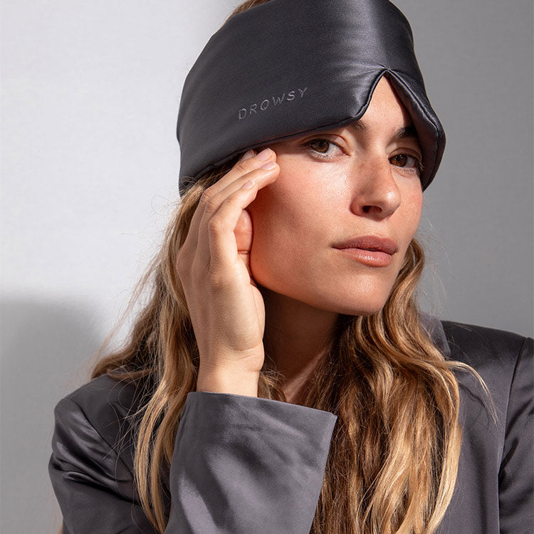 A woman with long, wavy blonde hair wears a dark grey silk sleep mask with DROWSY embroidered on it and a matching robe, looking thoughtfully at the camera against a neutral background.