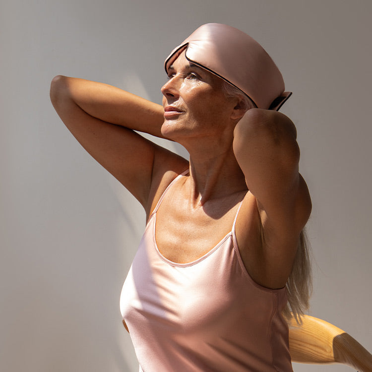 A woman in a pale pink satin outfit and matching sleep mask sits on a wooden chair, arms raised behind her head, gazing upward in soft natural light.