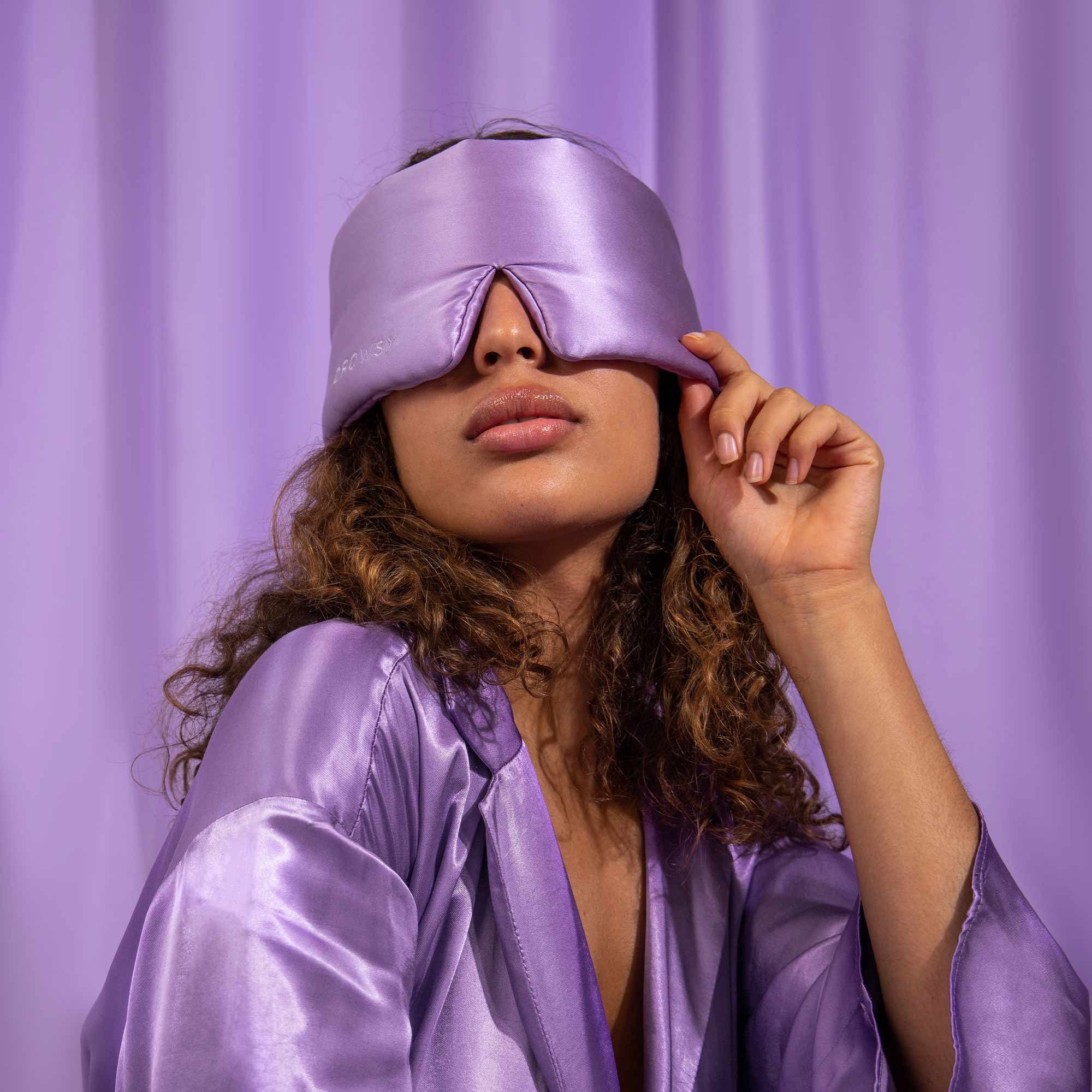 A woman poses in front of a lavender curtain, wearing the Bundle - Lavender Haze: a coordinating lavender satin robe and luxury silk mask, gently lifting the edge of the mask with one hand.