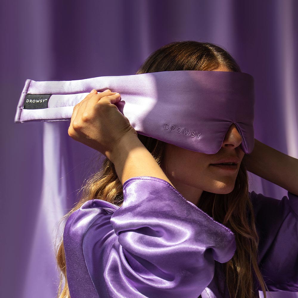 A woman wearing a shiny lavender robe adjusts a matching lavender sleep mask over her eyes. The mask has the word DROWSY embroidered on it. The background is also lavender.