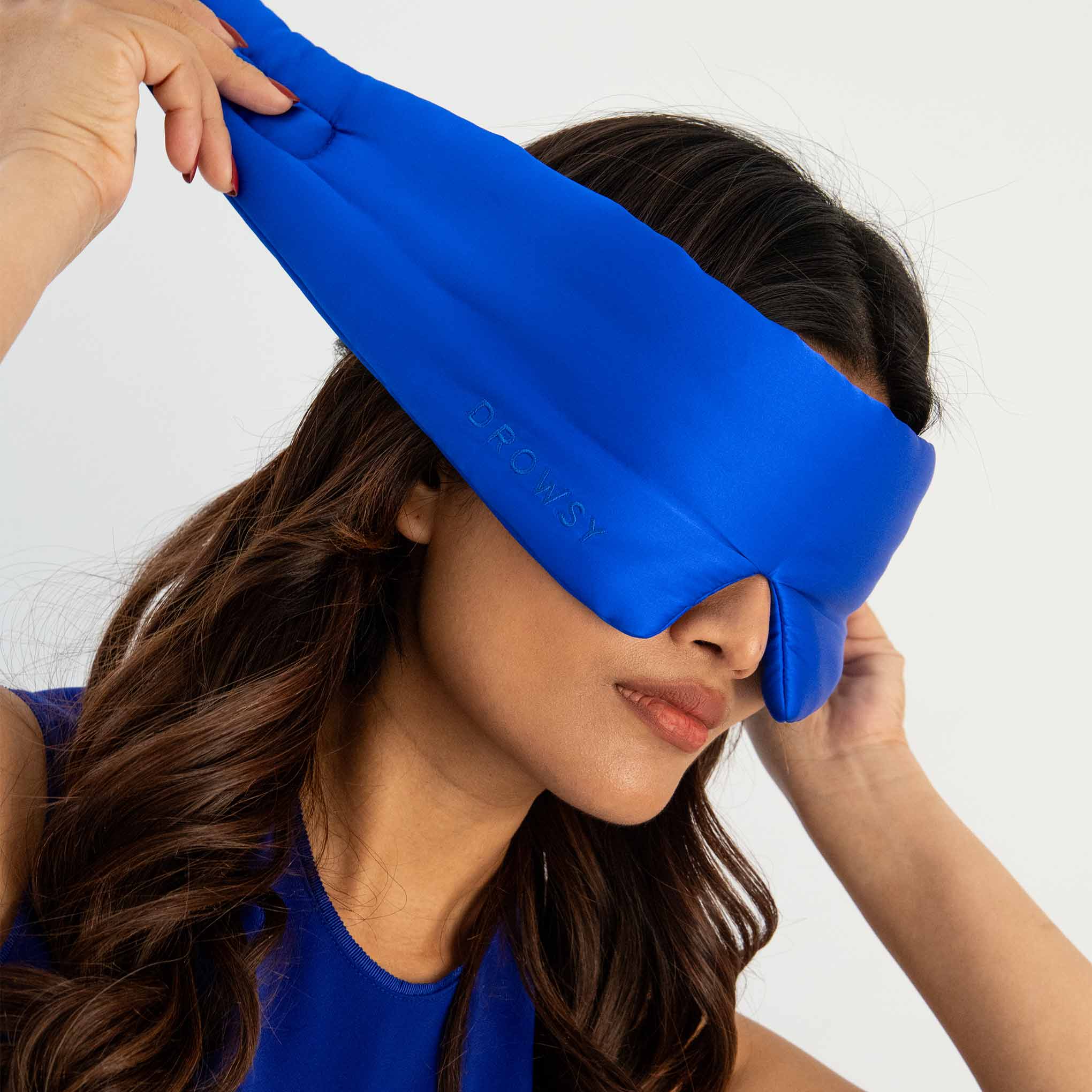 A woman with long brown hair puts on a bright blue sleep mask that covers her eyes. She is wearing a matching blue top and is adjusting the mask with one hand. The background is plain white.
