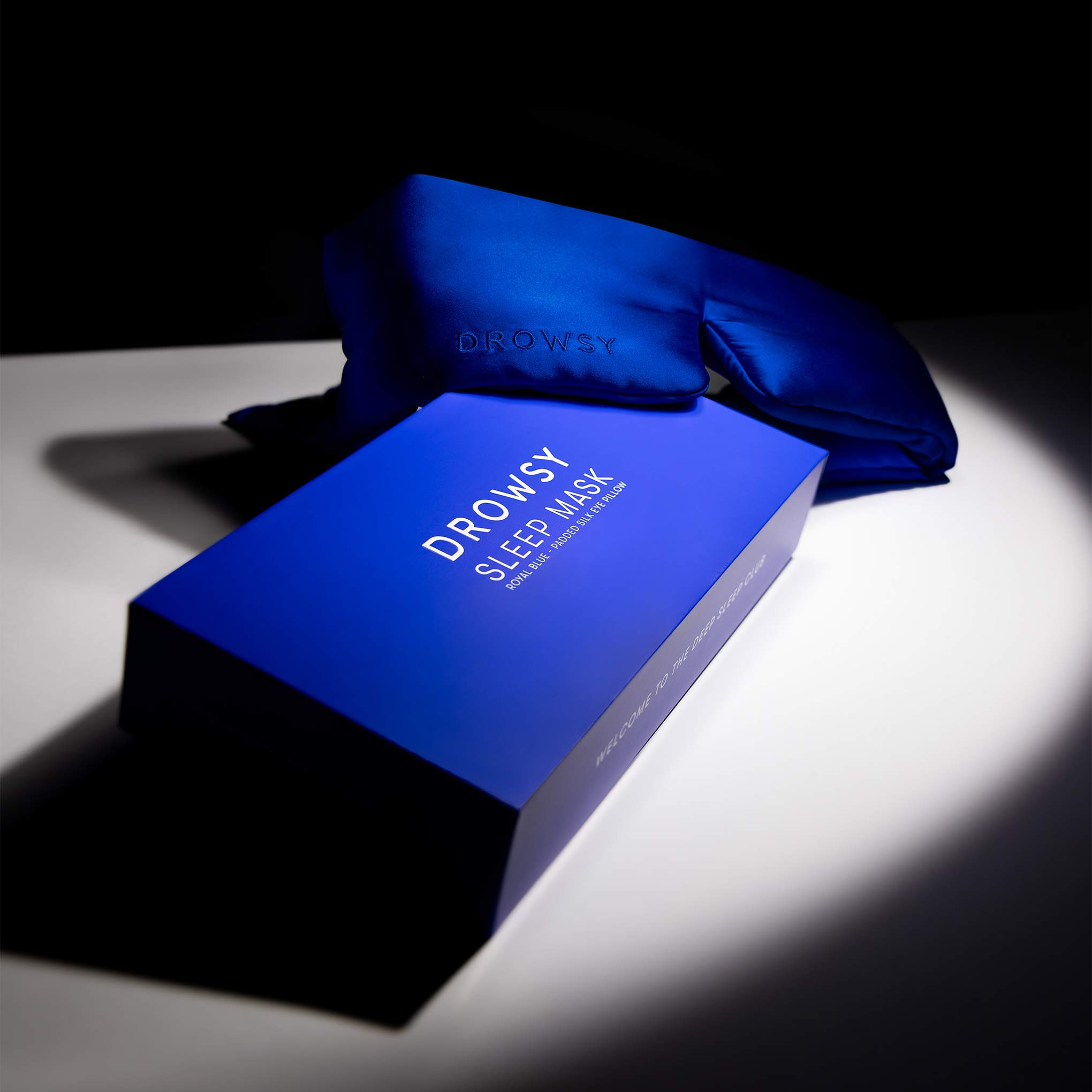 A royal blue box labeled Royal Blue sits on a white surface next to a matching silk eye mask, both illuminated by a spotlight against a dark background—perfect for luxurious beauty sleep.