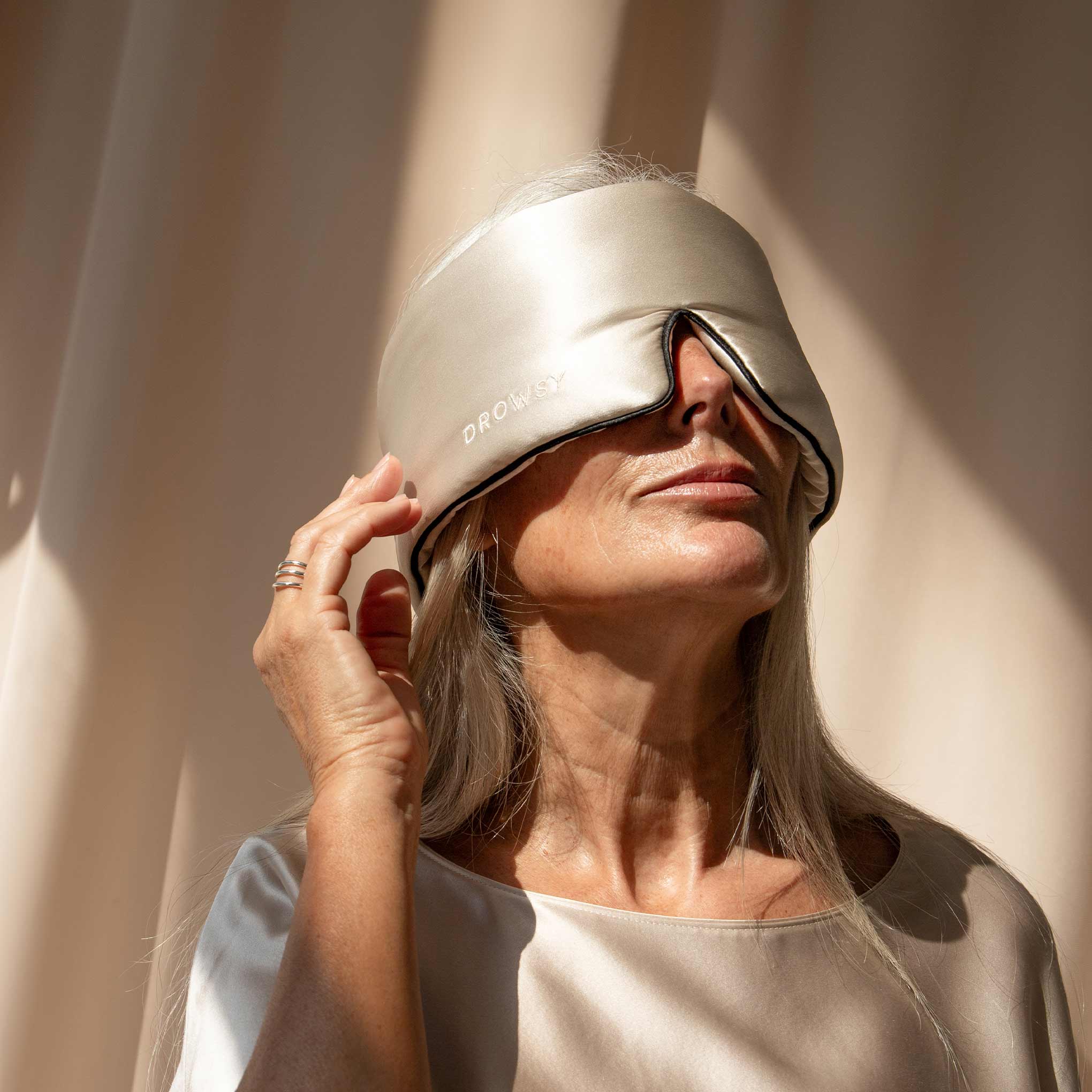 A woman with long gray hair stands in soft natural light, gently touching an oversized, shiny Dusty Gold Pipe Dreams Edition mulberry silk eye mask labeled DROWSY.