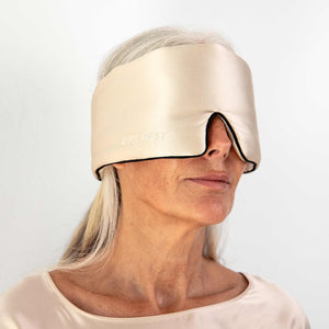 A woman with long gray hair wears a large, beige silk sleep mask labeled Dusty Gold Pipe Dreams Edition that covers her eyes and most of her forehead, standing against a plain white background.