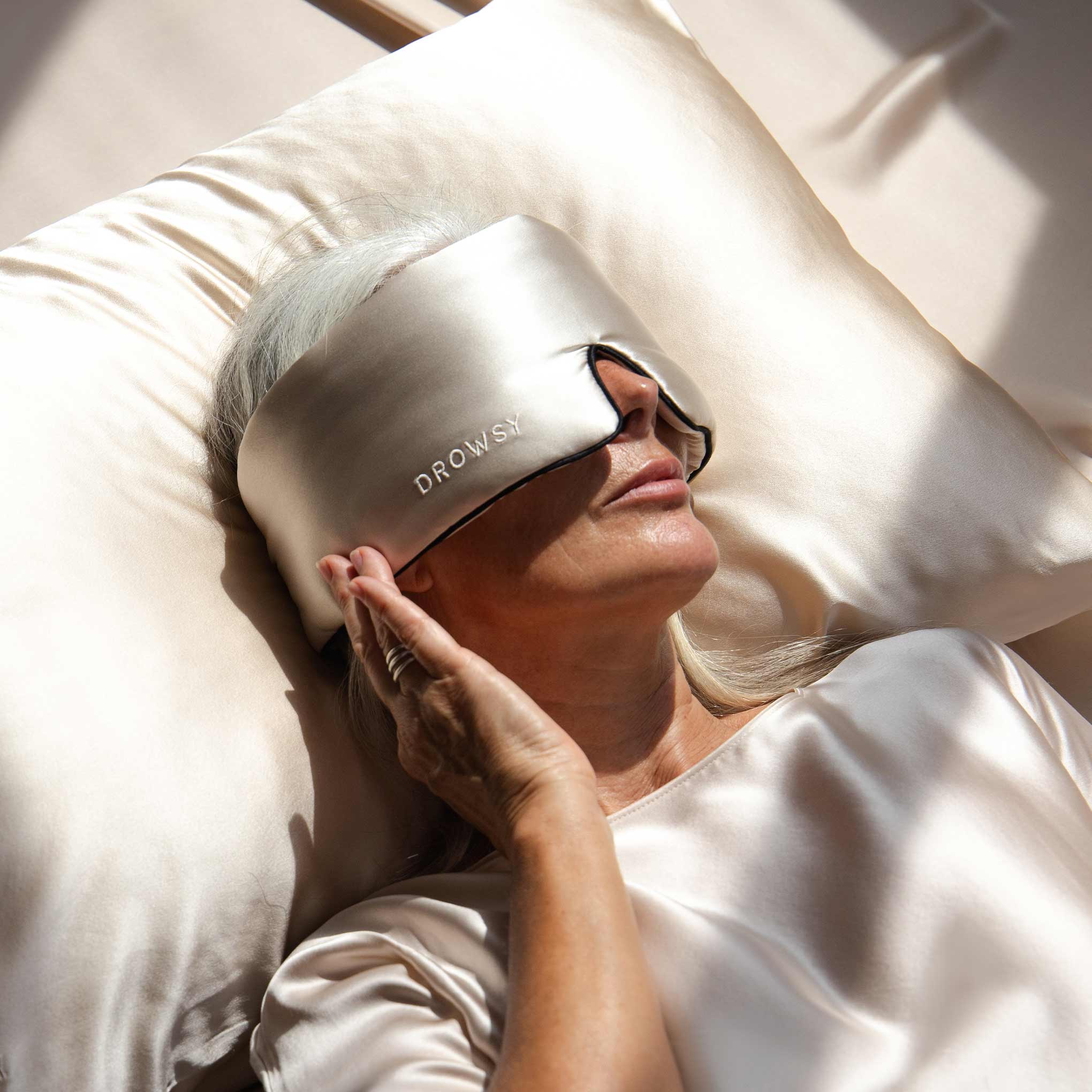 An older woman with gray hair lies on a satin pillow, wearing the Dusty Gold Pipe Dreams Edition mulberry silk eye mask labeled DROWSY and a matching satin top, gently touching the mask as soft light falls across her.