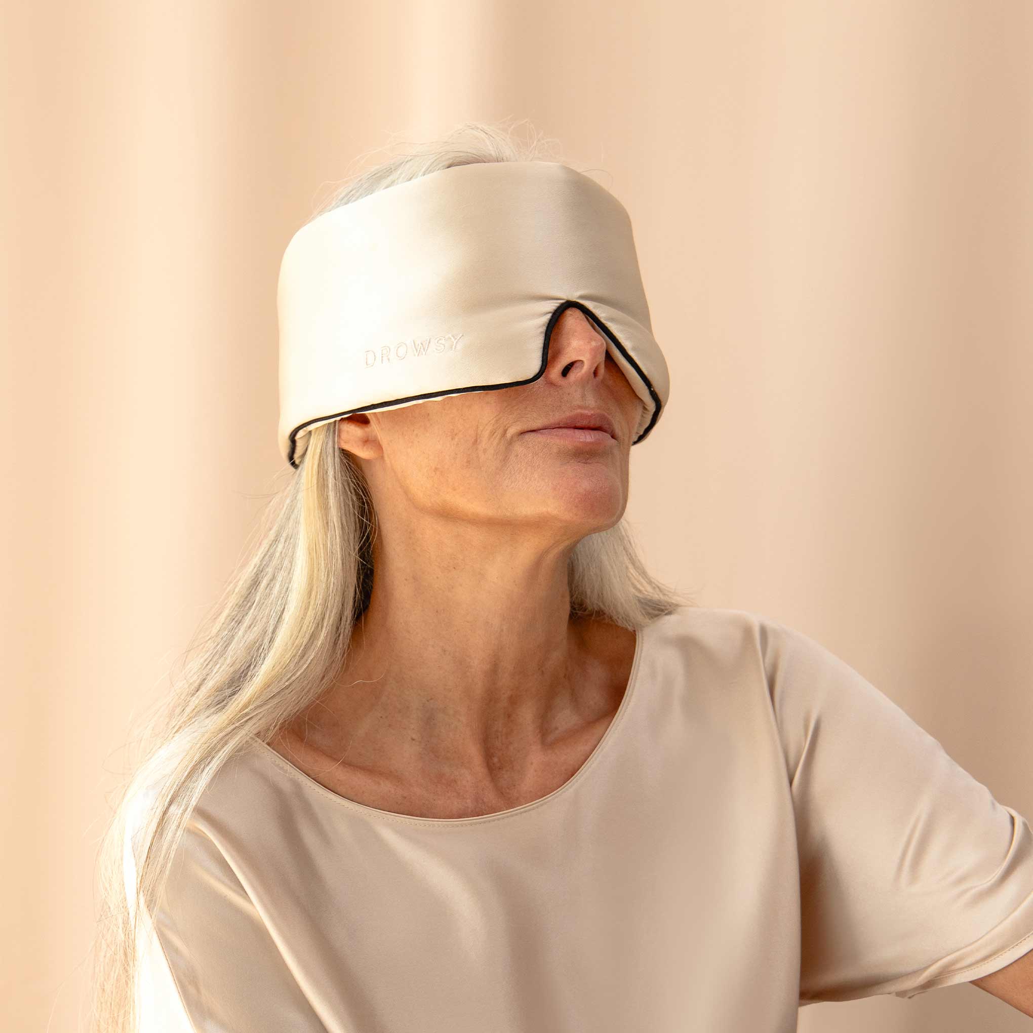 An older woman with long gray hair wears a large, beige Dusty Gold Pipe Dreams Edition mulberry silk eye mask with DROWSY visible, plus a matching shirt. She sits against a neutral background.