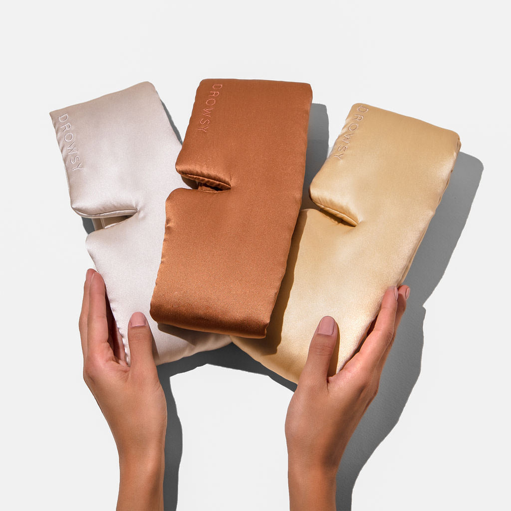 Two hands hold the Besties Pack—three silky sleep masks in ivory, copper, and gold—against a white background. Each mask features “DROWSY” embroidered on it.