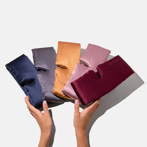 Two hands display the Family Pack: five SLOWSY silk eye masks in navy blue, gray, gold, pink, and burgundy, fanned out against a white background. Each mask features the embroidered SLOWSY brand name.