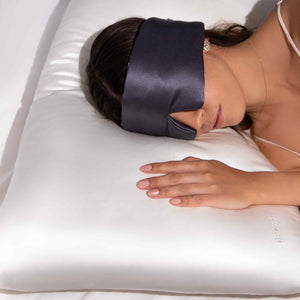 A woman rests on her side with one hand on a white Rajah Cushion Cover by drowsysleepco, wearing a black silk sleep mask and a white strap top. The pillow shows the PILLOW text partially visible.