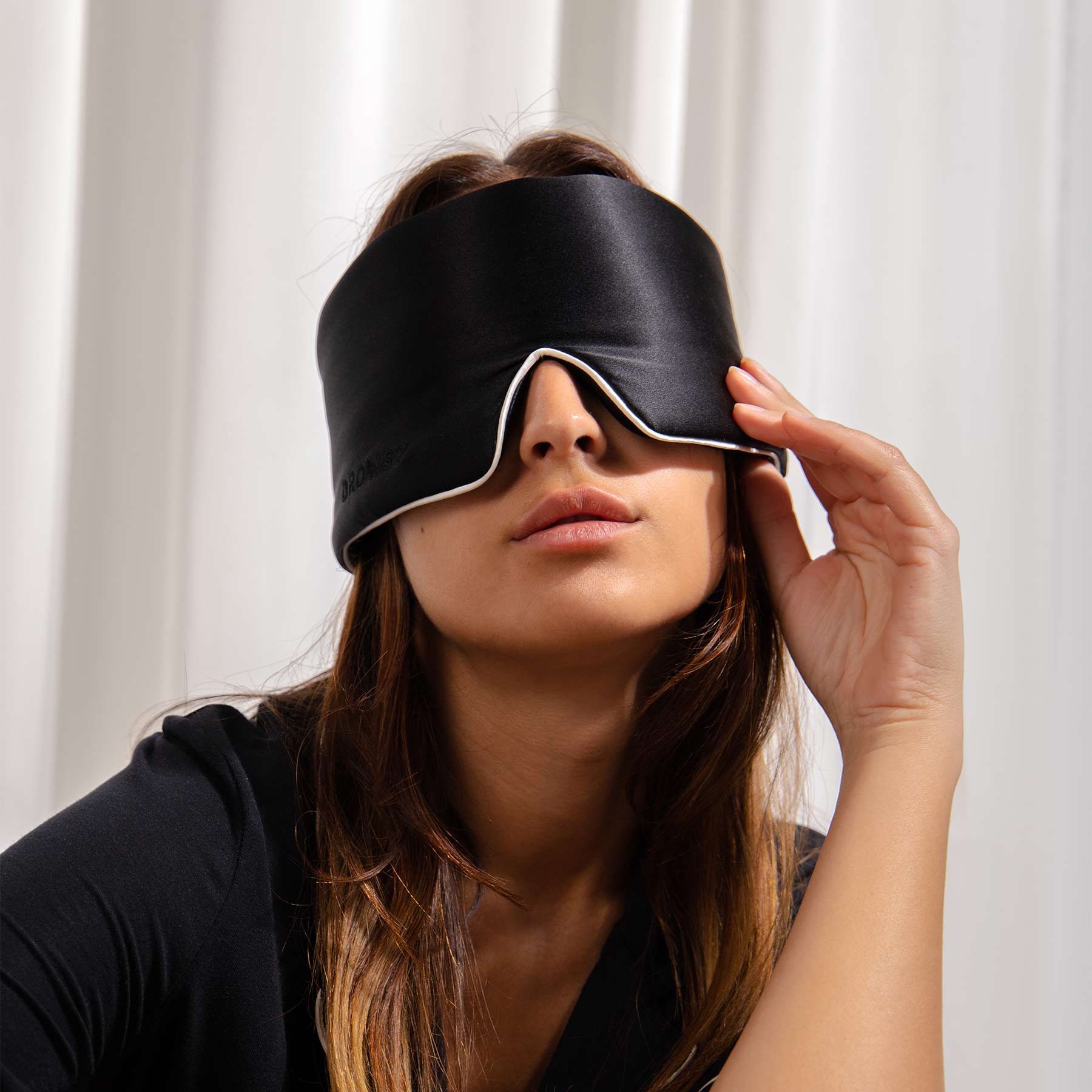 A woman with long brown hair wears the Black Jade - Pipe Dreams Edition, a large black silk sleep mask, and touches its edge while sitting in front of white curtains.