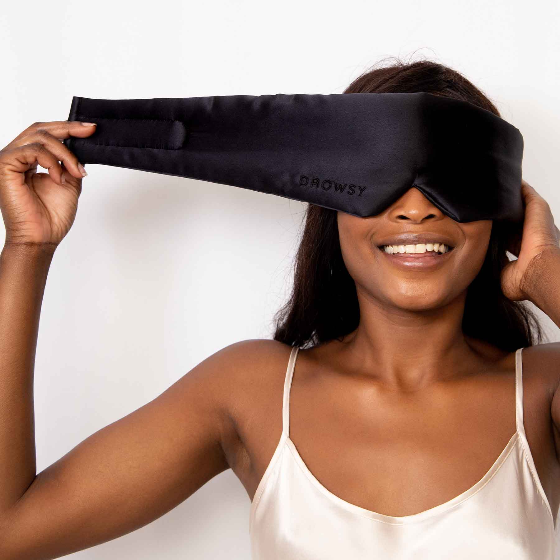 A woman wearing a satin sleep mask and smiling, with one hand adjusting the mask. She is dressed in a light-colored camisole against a plain white background.