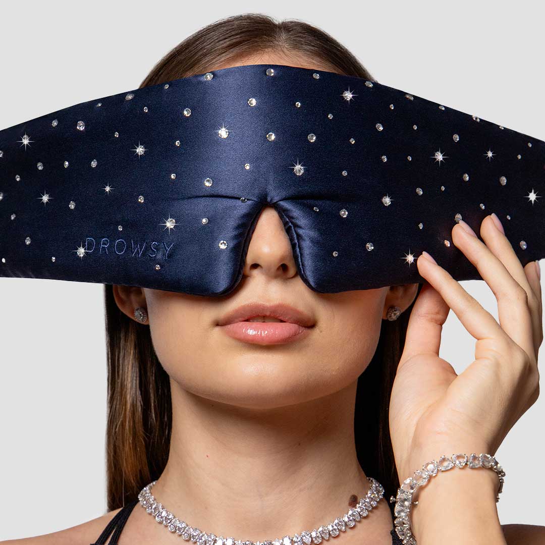 A woman wearing a navy blue, star-studded sleep mask covering her eyes and upper face, holding the sides of the mask. She is adorned with a diamond necklace and bracelet.