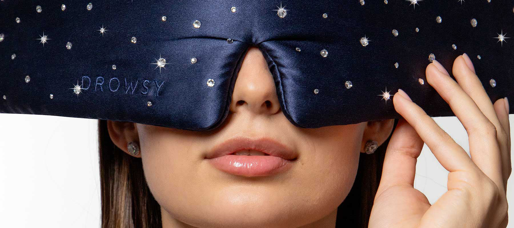 A woman wears a navy blue, jeweled sleep mask that covers her eyes and nose; the mask has DROWSY embroidered on it. She is holding one side of the mask and her lips are slightly parted.