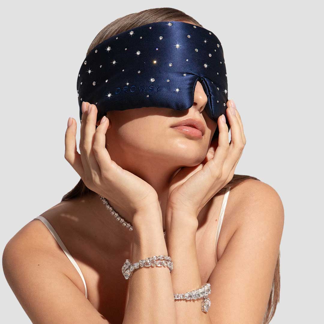 A woman wears the Drowsy Silk Eye Mask Crystals by Swarovski®, tilts her head back, touches her face, and accessorizes with Swarovski crystal bracelets, a necklace, and a white camisole.