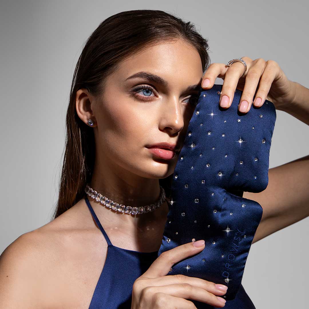 A woman in a blue halter dress holds the Drowsy Silk Eye Mask Crystals by Swarovski® to her face. She accessorizes with a crystal choker, earrings, and rings, her straight brown hair tucked neatly behind her ear.
