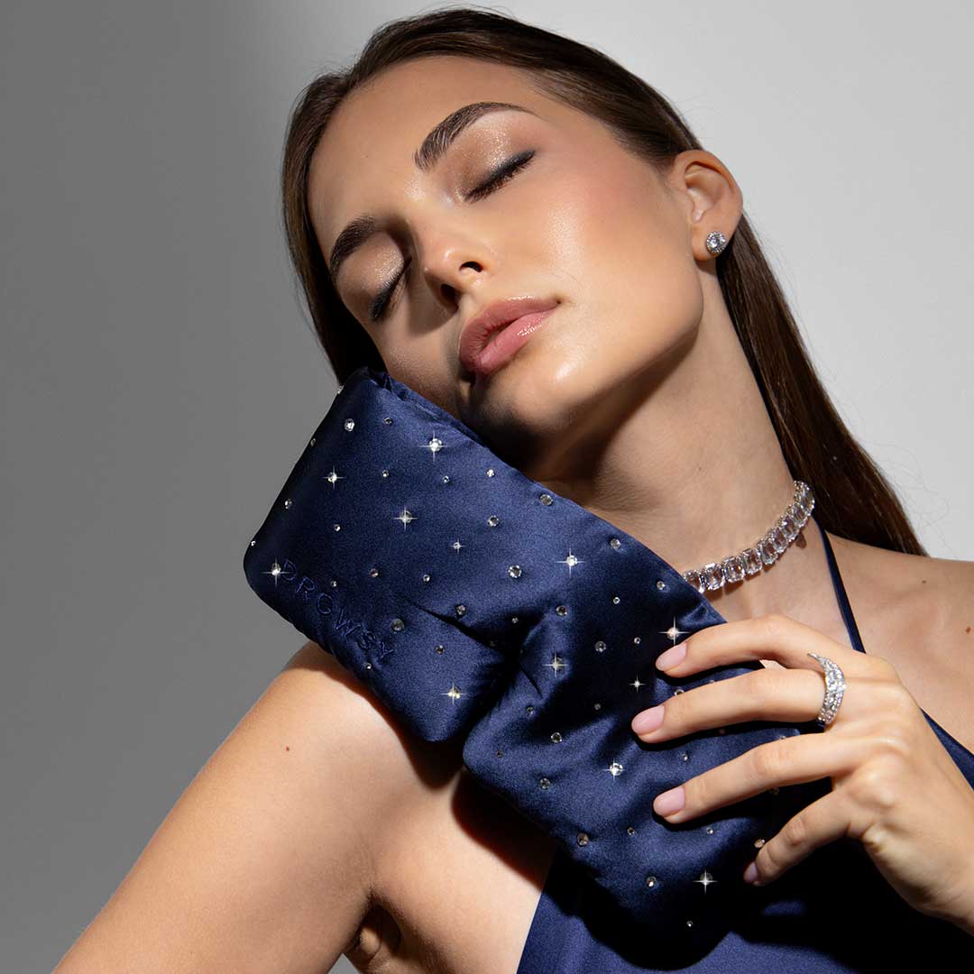 A woman with long brown hair and closed eyes rests the Drowsy Silk Eye Mask Crystals by Swarovski® on her cheek. Wearing a blue dress, sparkling necklace, and ring, she appears relaxed and serene.