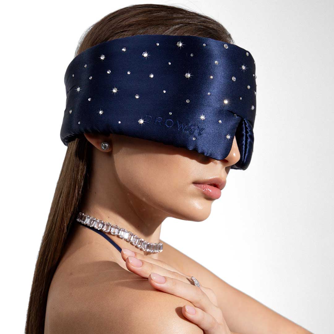 A woman wearing the Drowsy Silk Eye Mask Crystals by Swarovski® and a sparkly necklace poses with her hand on her shoulder against a white background.