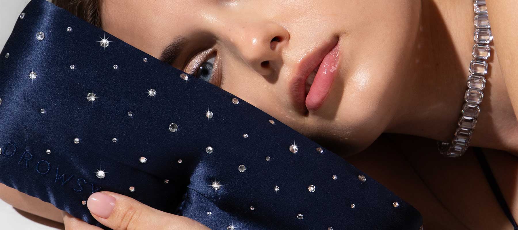 A woman with glossy skin and a crystal necklace rests her face on a navy blue sleep mask decorated with sparkling rhinestones.