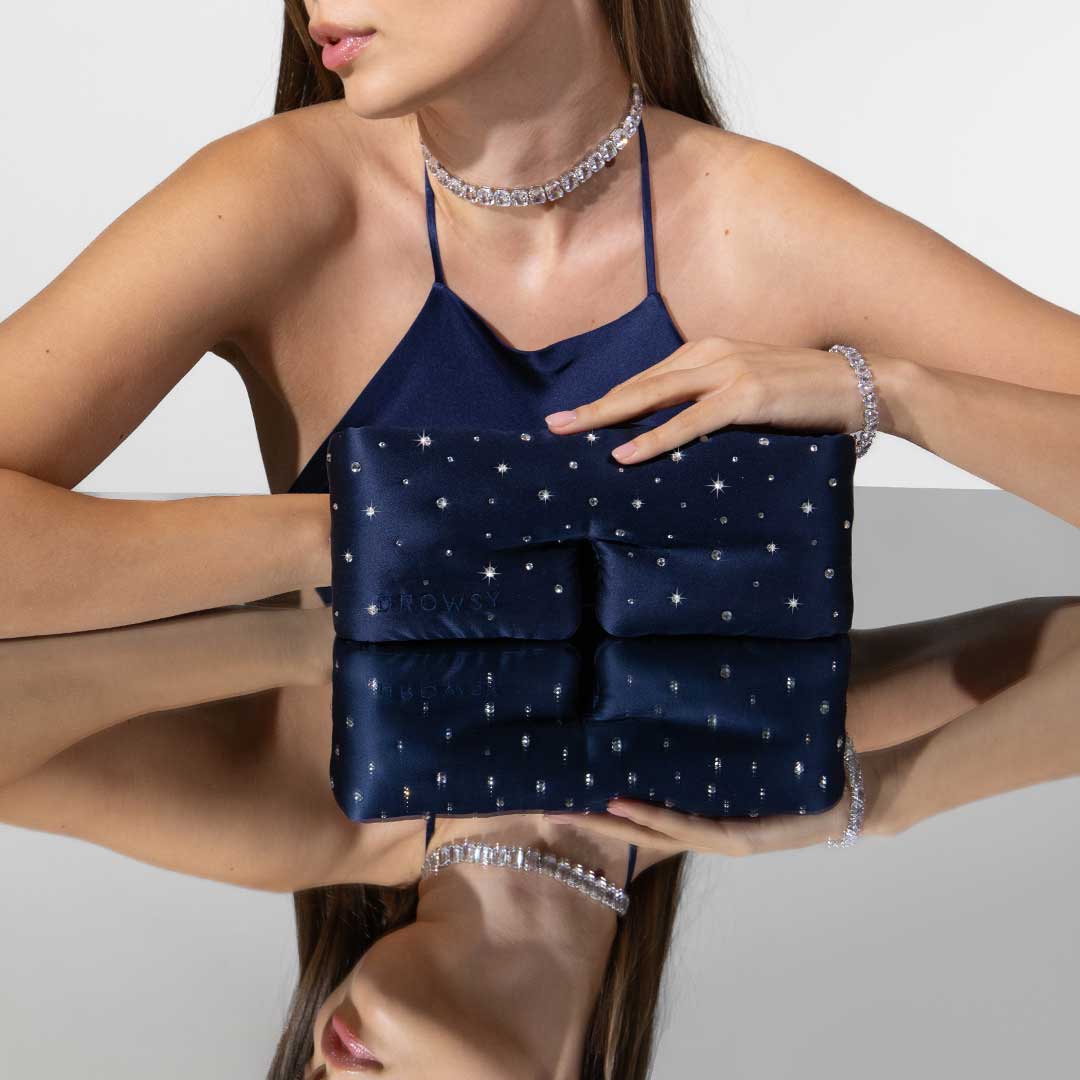 A woman in a navy halter top with a rhinestone choker and bracelet holds the Drowsy Silk Eye Mask Crystals by Swarovski®, its shimmering surface reflected on a mirror. Her face is partially out of frame.
