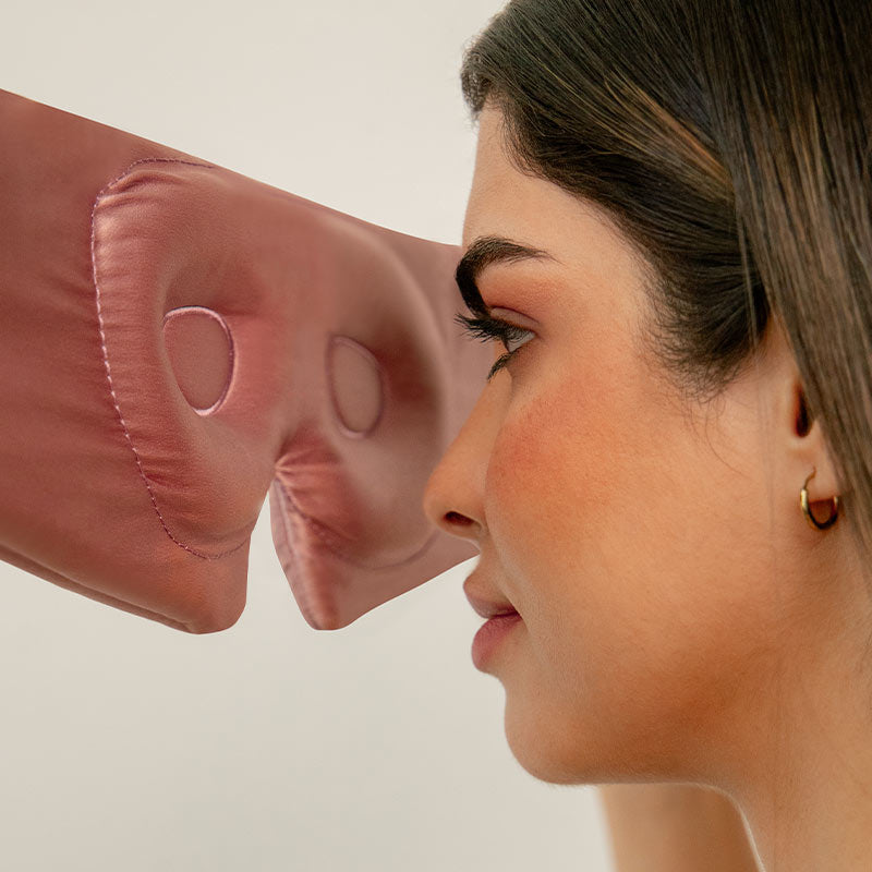 A woman faces a soft, pink, padded object with two eye holes, resembling a mask or cushion, positioned near her eyes against a plain background.