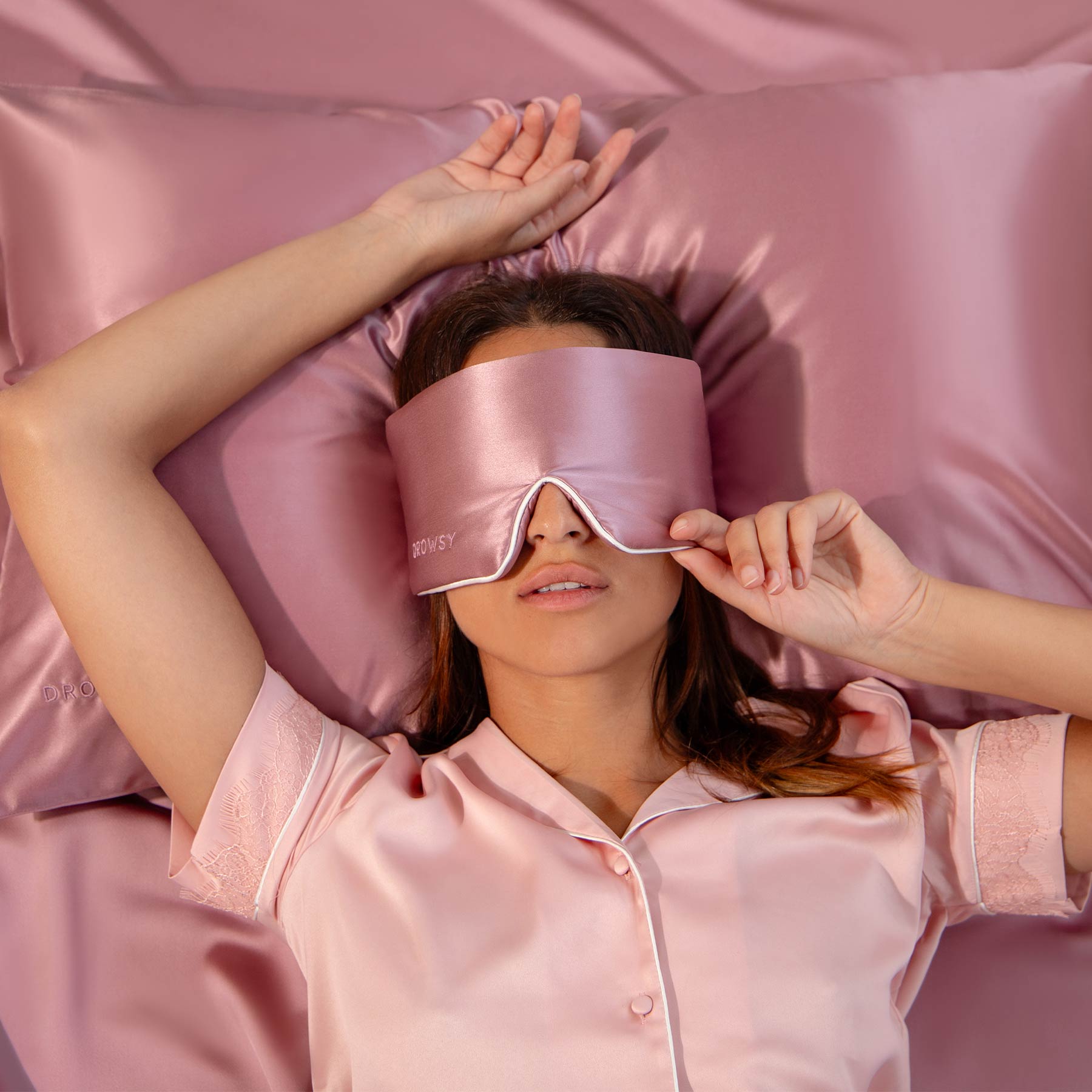 A woman relaxes on a bed dressed in Damask Rose - Pipe Dreams Edition pink satin sheets and pajamas, wearing a large adjustable mulberry silk sleep mask, one hand on her head, the other lightly touching the luxurious mask.