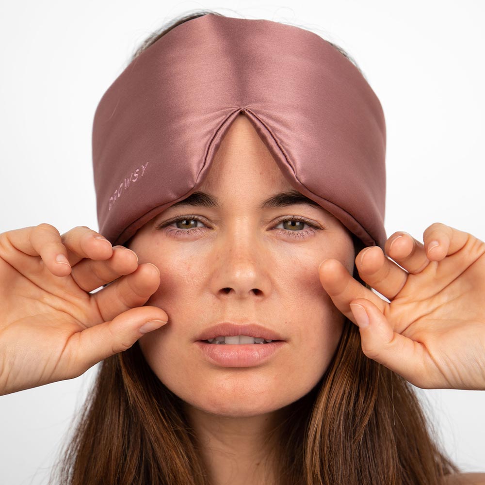 A woman with long brown hair wears the Damask Rose silk eye mask pushed up on her forehead, gently touching its sides. She gazes at the camera with a calm expression, capturing effortless elegance and beauty sleep style.